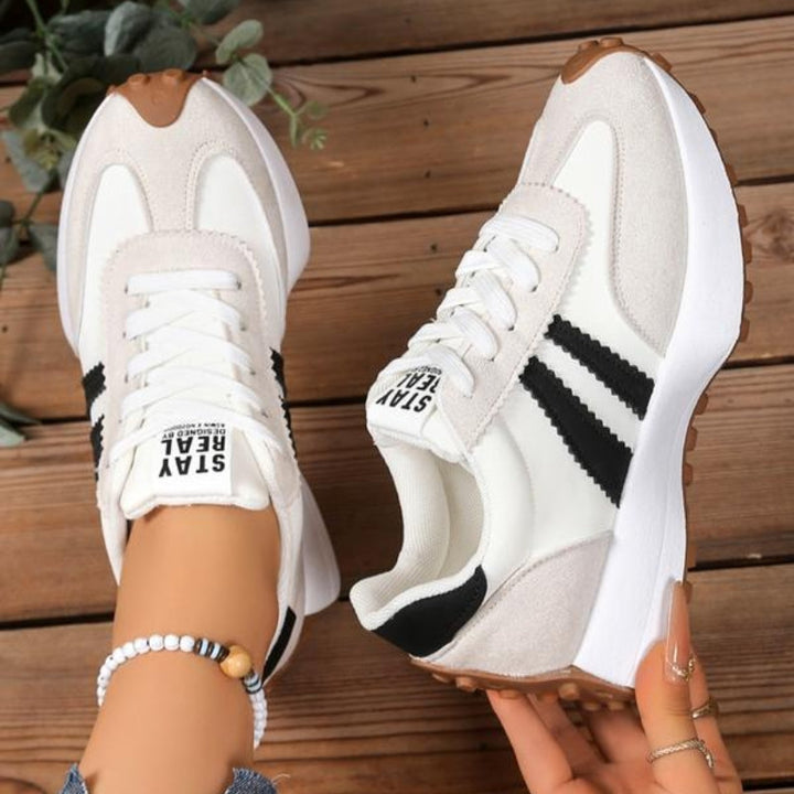 Weiß-beige Sneaker mit schwarzen Streifen, einer getragen, der andere in der Hand, auf Holzboden 10