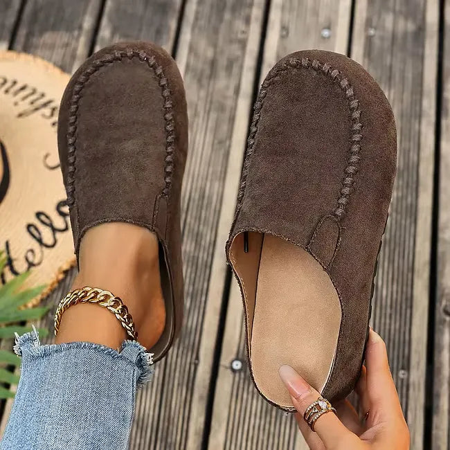Dunkelbraune Wildleder-Slipper von oben, ein Schuh getragen, der andere mit sichtbarem hellem Fußbett in der Hand gehalten. 3