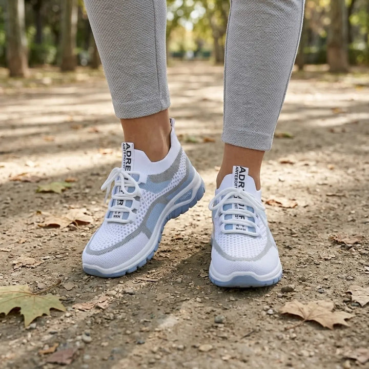 Weiß-hellblaue Mesh-Sneaker an den Füßen einer Trägerin im Park, kombiniert mit grauer Hose, auf Waldboden mit Herbstlaub. 2