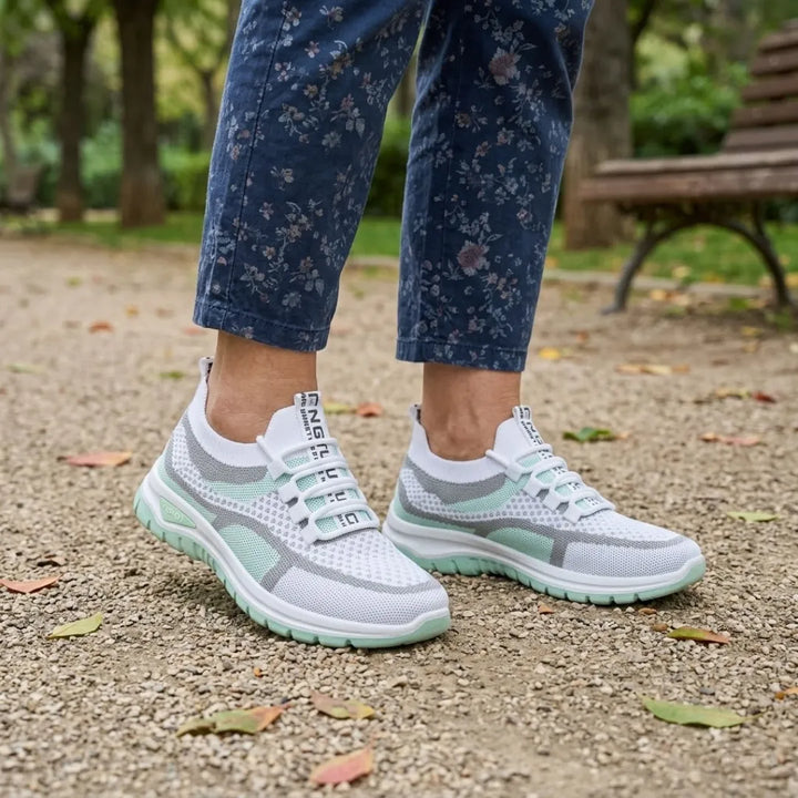 Weiß-mintgrüne Mesh-Sneaker an den Füßen einer Trägerin mit geblümter blauer Hose im Park vor einer Holzbank. 11
