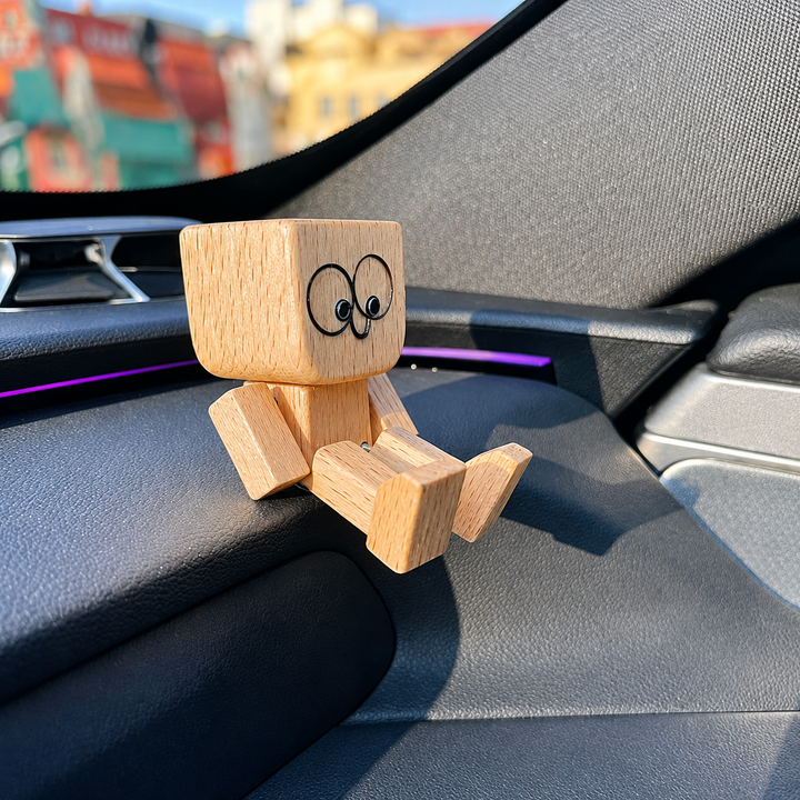Kleine Holzfigur mit Kulleraugen sitzt seitlich fotografiert im Auto auf der Mittelkonsole, im Hintergrund bunte Häuser durch die Windschutzscheibe. 2