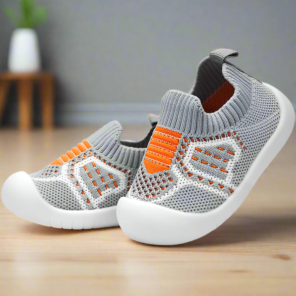 Paar graue Strickschuhe mit orangen Details, weißer Gummikappe und Fersenschlaufe, schräg auf Holzboden fotografiert. 16