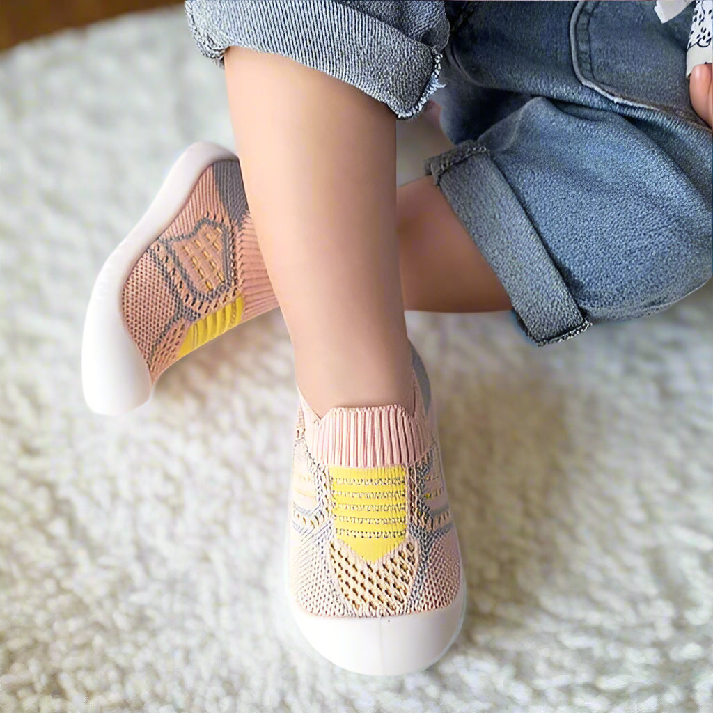 Kinderfüße in rosa Strickschuhen mit gelben Streifen und Netzeinsatz auf einem weißen Teppich, ein Schuh schräg aufgestellt. 13