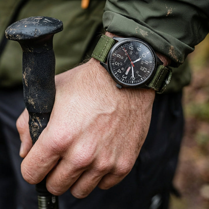 Nahaufnahme am Handgelenk eines Wanderers mit Trekkingstock, die Uhr mit grünem Stoffarmband zu schmutziger Outdoorjacke. 2