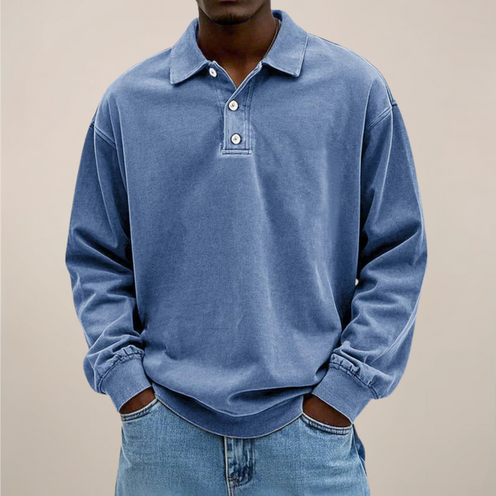 Mann trägt das lockere Langarm-Poloshirt in Mittelblau, kombiniert mit heller Jeans und beide Hände in den Hosentaschen. 1