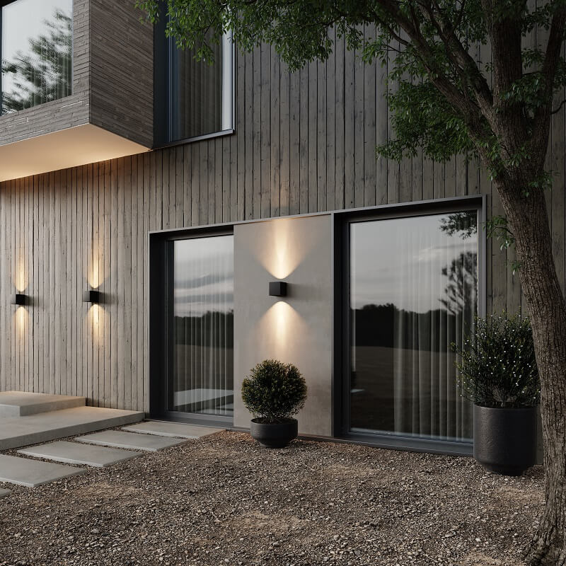 Mehrere schwarze Wandleuchten an einer dunklen Holzfassade mit Terrasse und Pflanzkübeln werfen abends Lichtkegel nach oben und unten. 3