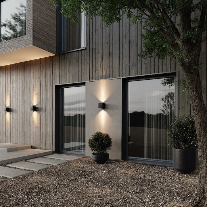 Mehrere schwarze Wandleuchten an einer dunklen Holzfassade mit Terrasse und Pflanzkübeln werfen abends Lichtkegel nach oben und unten. 3