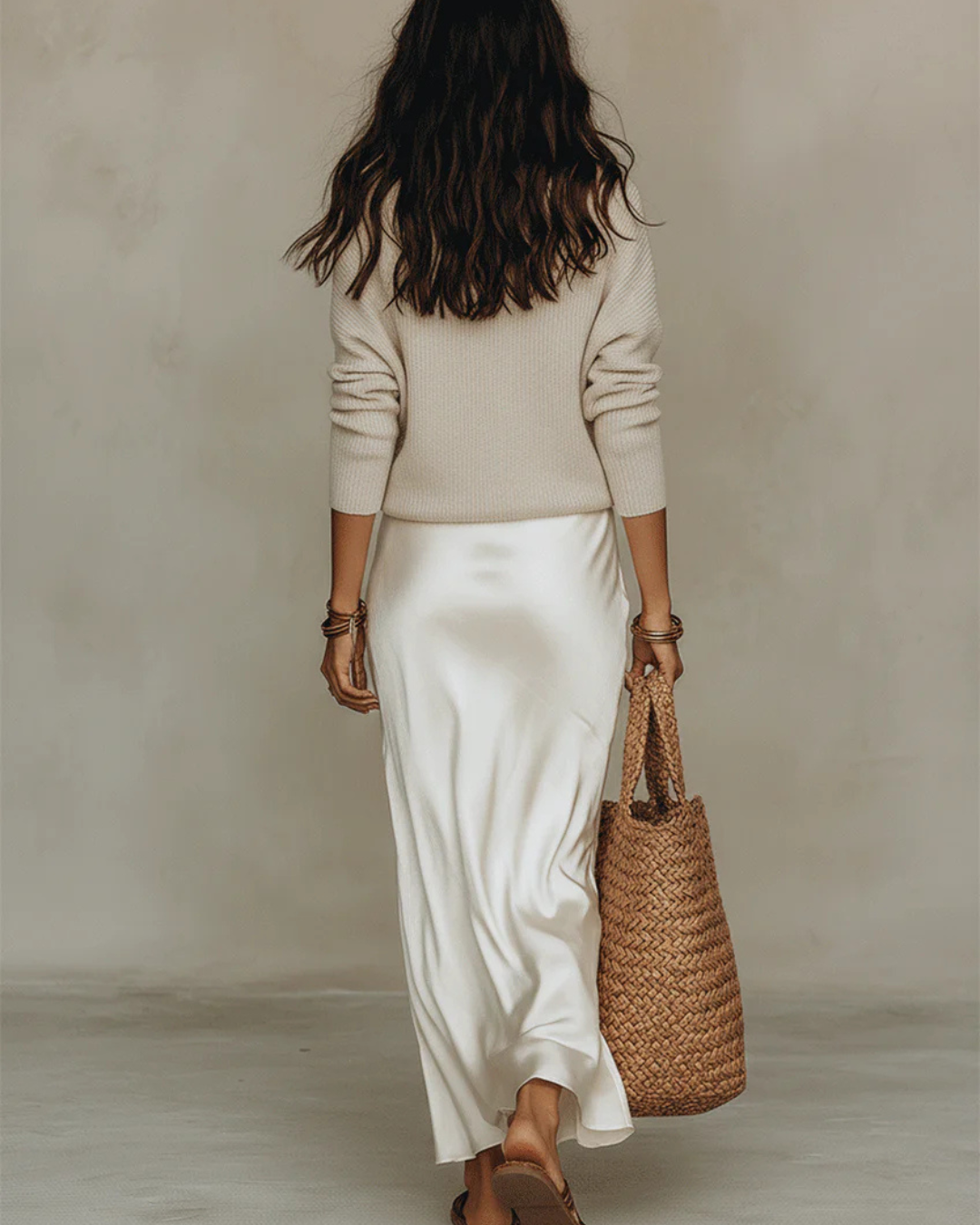 Rückansicht des Models in cremefarbenem Satinkleid und beigem Strickpullover, mit Korbtasche in der Hand 9