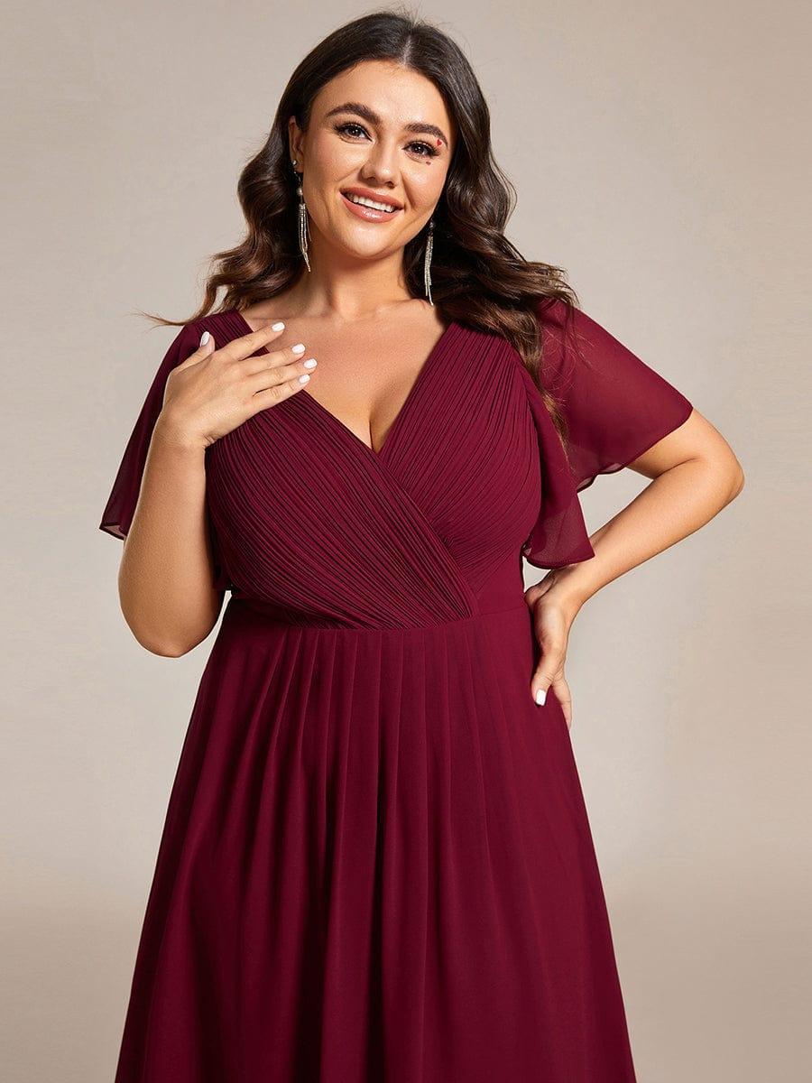 Oberkörper-Nahaufnahme des weinroten Kleids am Plus-Size-Model: gerafftes Wickeloberteil und Flatterärmel, eine Hand an der Brust. 51