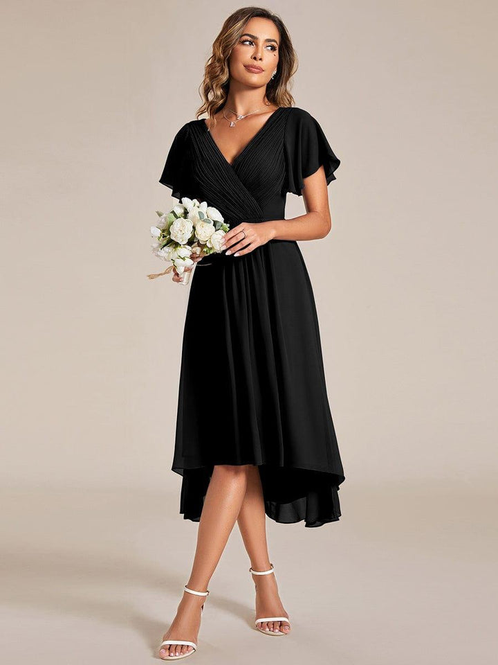 Model im schwarzen Chiffonkleid hält einen Strauß weißer Rosen, die Beine sind überkreuzt, der Saum verläuft vorne kürzer als hinten. 24
