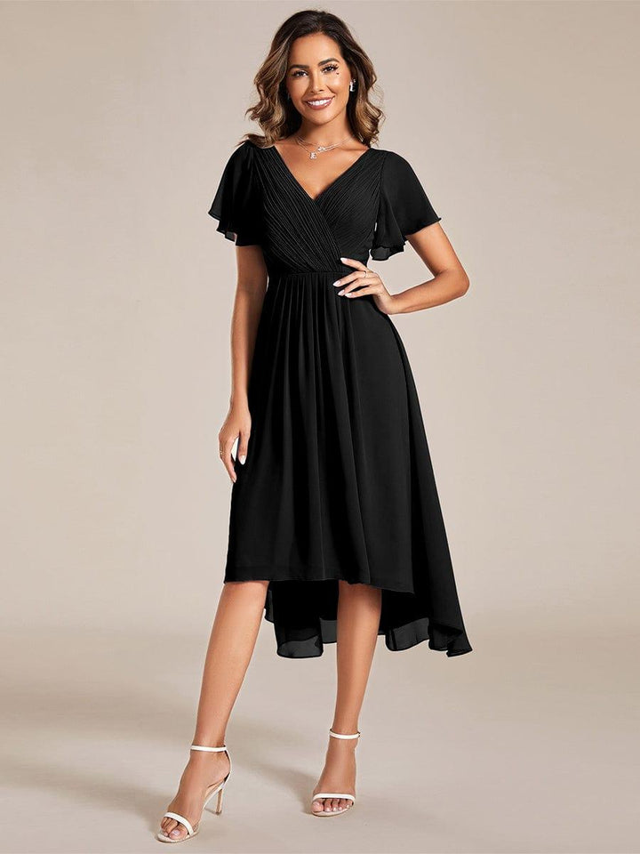 Frau in schwarzem Chiffonkleid mit V-Ausschnitt und Flatterärmeln, Hand in der Hüfte, Vorderansicht mit asymmetrischem Saum und weißen Sandaletten. 21