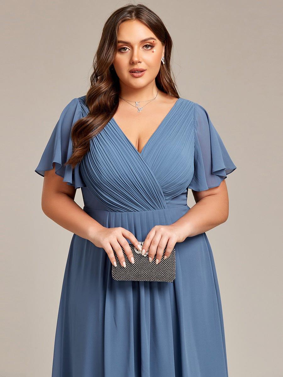 Nahaufnahme des gerafften V-Ausschnitts und der Flatterärmel in Staubblau, das Model hält eine schwarze Glitzer-Clutch. 43