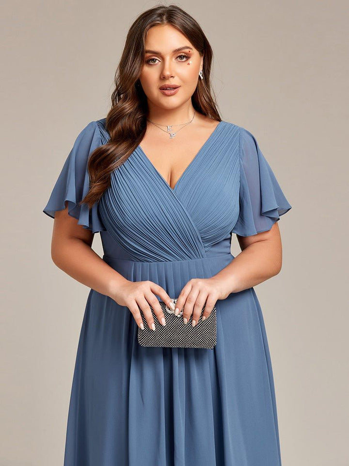 Nahaufnahme des gerafften V-Ausschnitts und der Flatterärmel in Staubblau, das Model hält eine schwarze Glitzer-Clutch. 43
