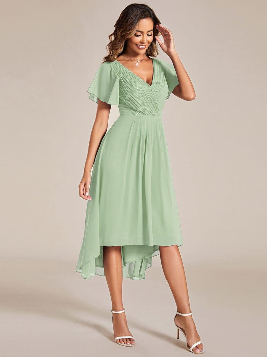Grünes Chiffonkleid am Model in Dreiviertelansicht, eine Hand am Kopf, mit gerafftem V-Ausschnitt, Flatterärmeln und vorne kürzerem Saum. 80