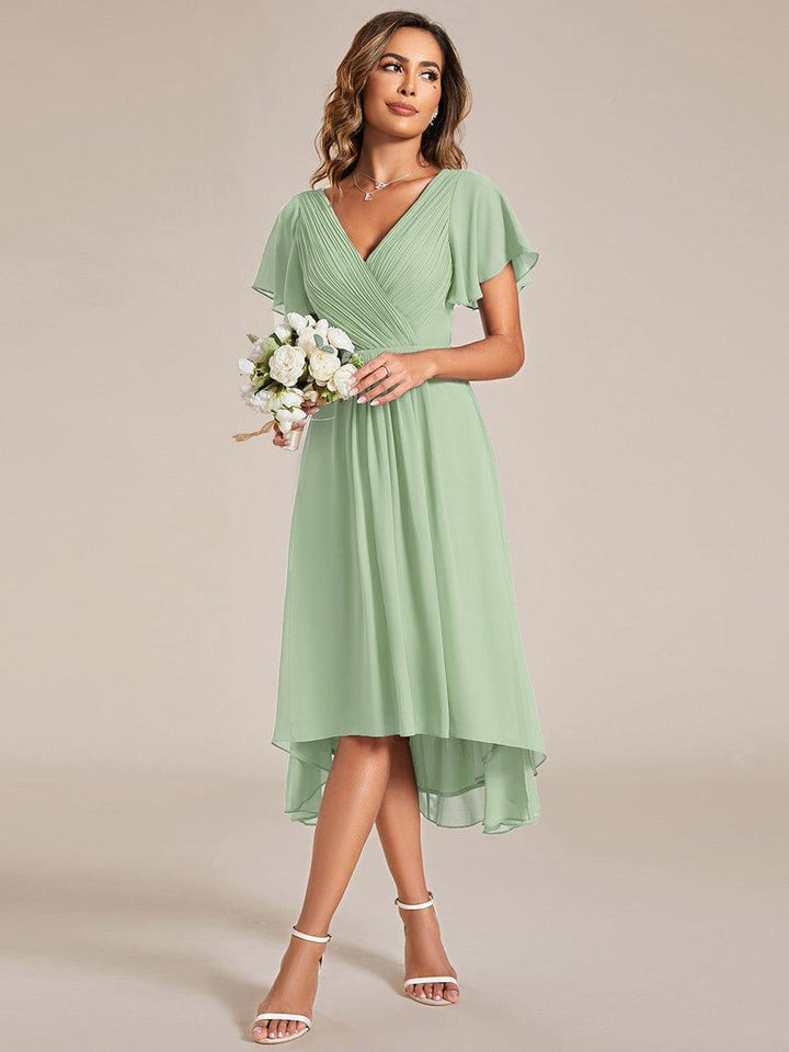 Model in grünem Chiffonkleid hält einen kleinen weißen Blumenstrauß; Wickeloberteil, Flatterärmel und asymmetrischer Saum sind zu sehen. 79