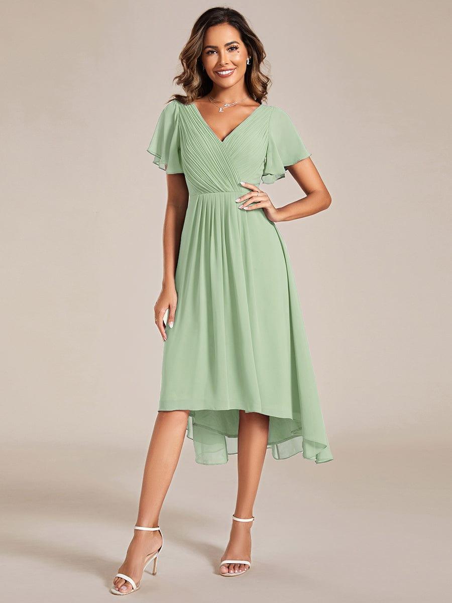 Salbeigrünes Chiffonkleid in Ganzkörperansicht am Model, mit Wickeloberteil, Flatterärmeln und Hoch-Tief-Saum, eine Hand an der Hüfte, dazu weiße Sandaletten. 76
