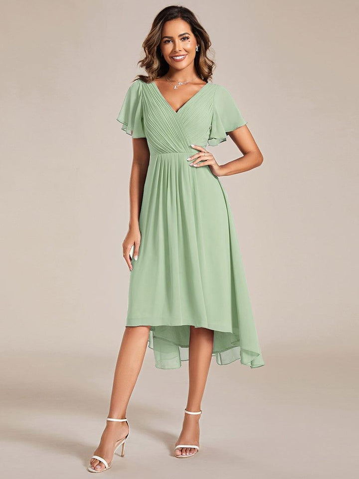 Salbeigrünes Chiffonkleid in Ganzkörperansicht am Model, mit Wickeloberteil, Flatterärmeln und Hoch-Tief-Saum, eine Hand an der Hüfte, dazu weiße Sandaletten. 76