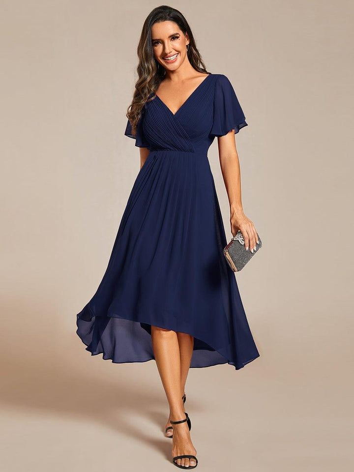 Frontansicht des Kleids in Marineblau mit plissiertem V-Ausschnitt, Flatterärmeln und asymmetrischem Saum, getragen mit glitzernder Clutch. 1