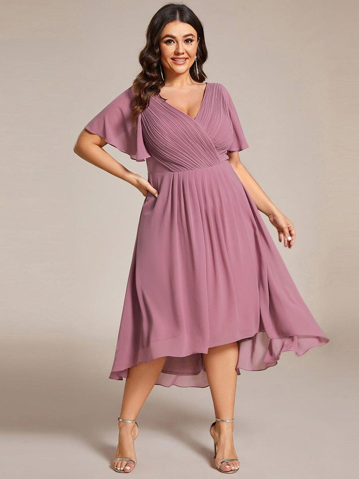 Curvy-Model in mauvefarbenem Chiffonkleid, eine Hand an der Hüfte, mit goldfarbenen Sandaletten; der Rock fällt vorne kürzer als hinten. 66