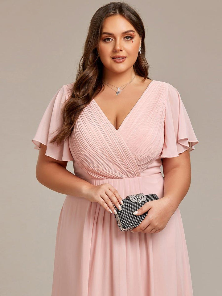 Nahaufnahme des rosafarbenen Oberteils mit gerafftem V-Ausschnitt und Flatterärmeln, das Curvy Model hält eine schwarze Glitzerclutch. 33
