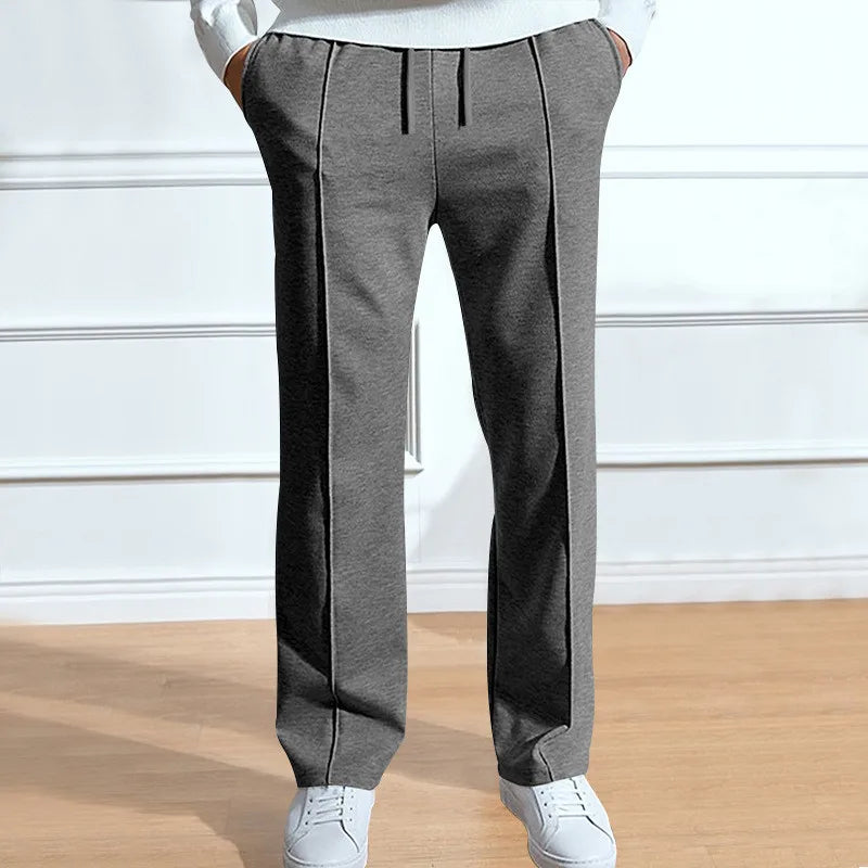 Graue Hose mit Kordelzug und Bügelfalten, getragen mit weißem Oberteil und weißen Sneakern, Hände in den Taschen. 2