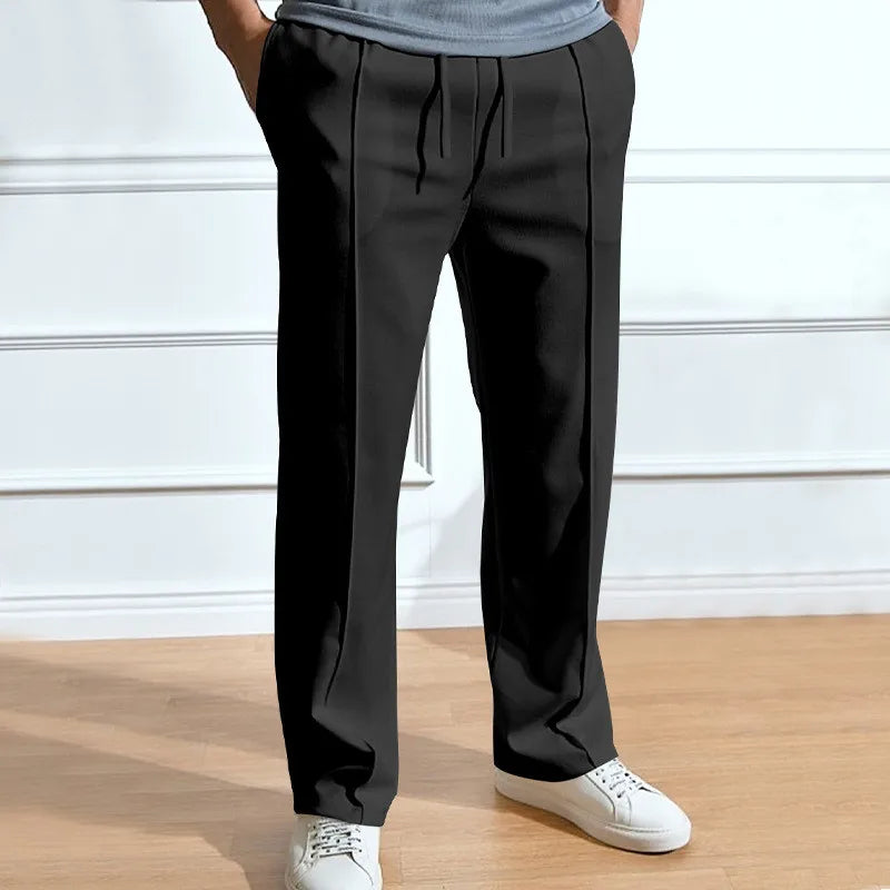 Schwarze Hose mit Kordelzug und Bügelfalten, getragen mit grauem Shirt und weißen Sneakern vor heller Wand. 3