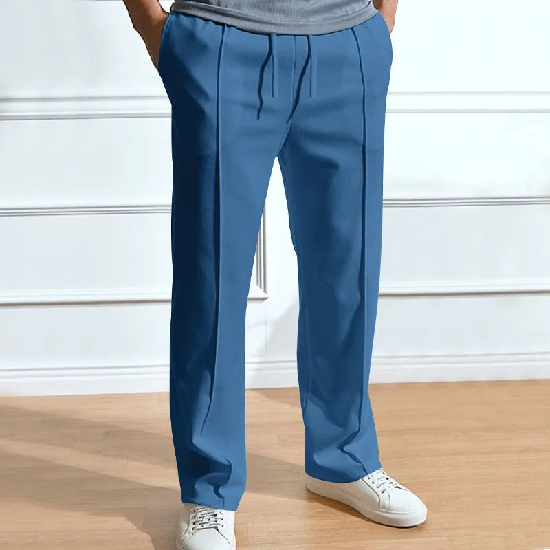 Blaue Hose mit Kordelzug und Bügelfalten, getragen mit grauem Shirt und weißen Sneakern vor heller Wand. 5