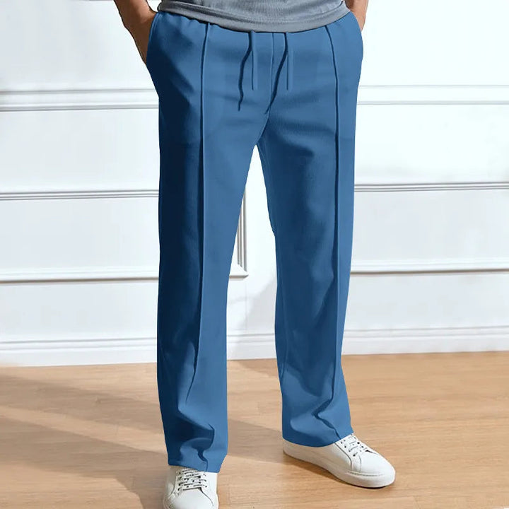 Blaue Hose mit Kordelzug und Bügelfalten, getragen mit grauem Shirt und weißen Sneakern vor heller Wand. 5