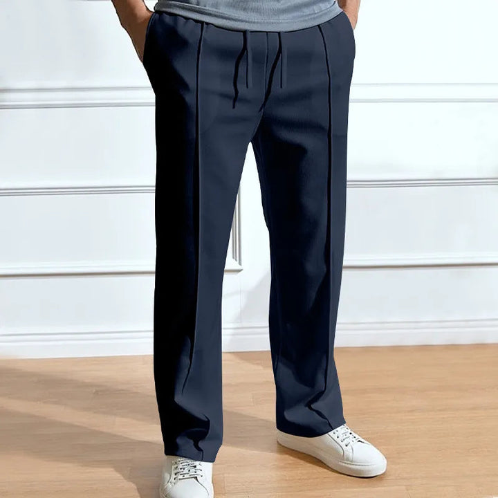 Dunkelblaue Hose mit Kordelzug und Bügelfalten, getragen mit grauem Shirt und weißen Sneakern vor heller Wand. 1