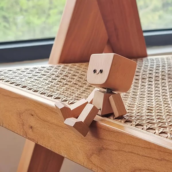 Holzfigur mit lächelndem Gesicht sitzt auf der Kante eines geflochtenen Holzstuhls, die Beine hängen über den Rand. 5