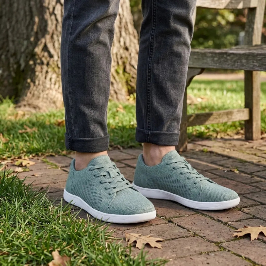 Person in dunkler Jeans mit hochgekrempelten Hosenbeinen trägt graugrüne Mesh-Sneaker mit weißer Sohle auf einem Backsteinweg im Park. 29