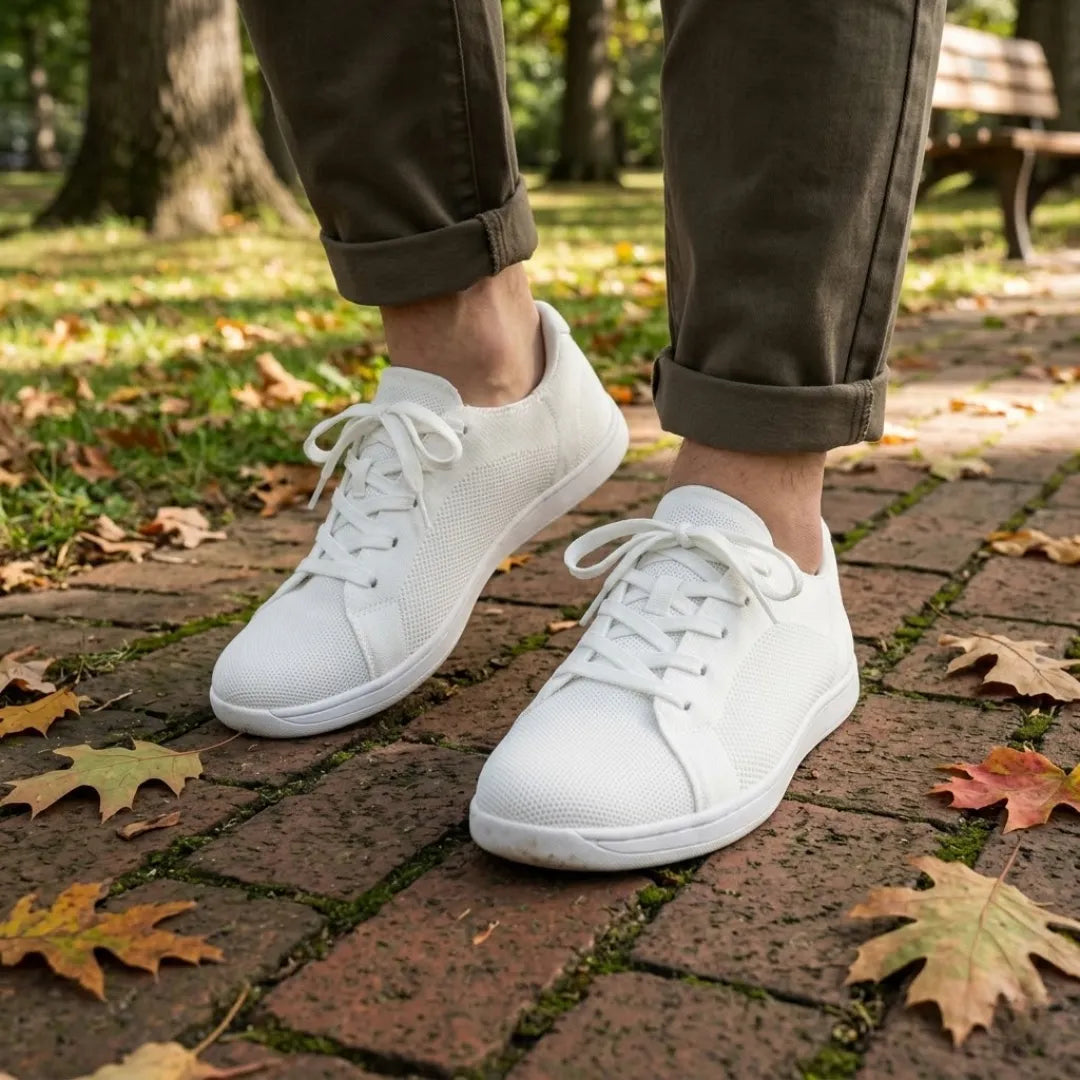 Person in olivgrüner Hose mit hochgekrempelten Hosenbeinen trägt weiße Mesh-Sneaker auf einem Backsteinweg mit Herbstlaub im Park. 38