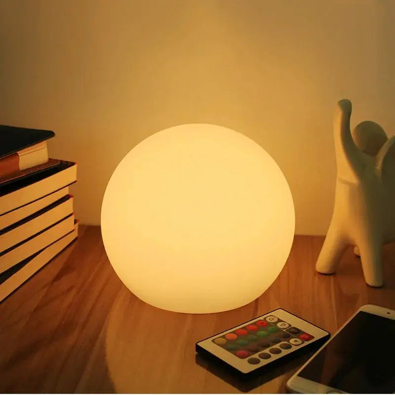 Warm leuchtende Kugellampe auf einem Holztisch, daneben Fernbedienung mit bunten Tasten, Smartphone, Bücherstapel und weiße Keramikfigur. 1