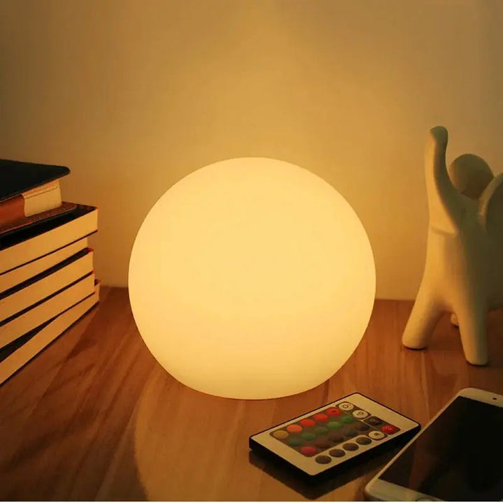 Warm leuchtende Kugellampe auf einem Holztisch, daneben Fernbedienung mit bunten Tasten, Smartphone, Bücherstapel und weiße Keramikfigur. 1