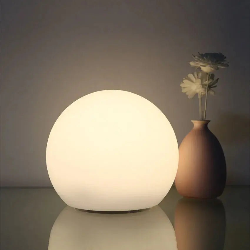Weich warmweiß leuchtende Kugellampe neben einer schlichten Vase mit Blumen, die sich auf der glatten Fläche spiegeln. 2