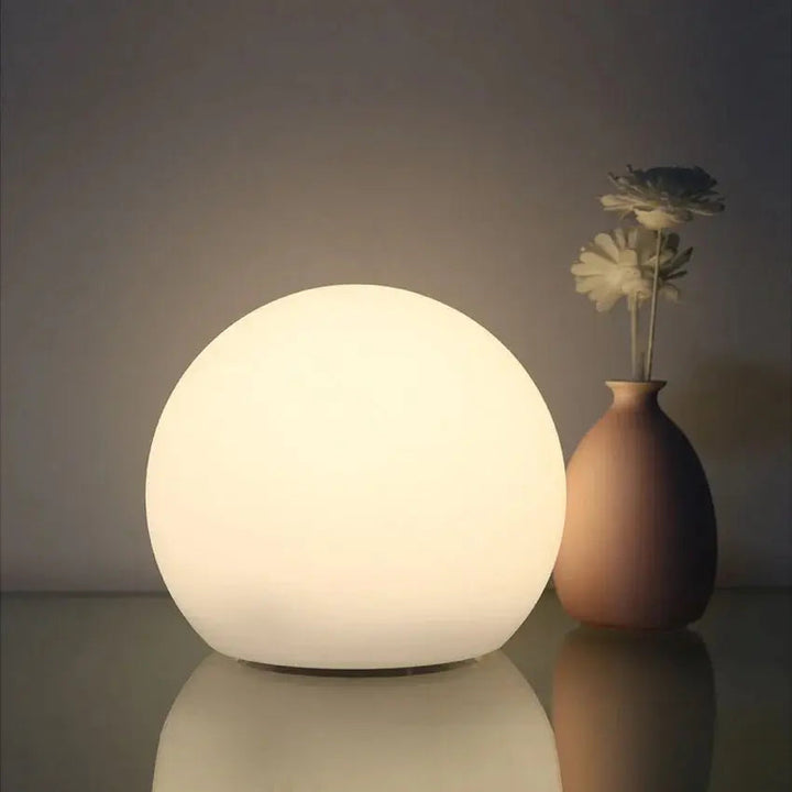 Weich warmweiß leuchtende Kugellampe neben einer schlichten Vase mit Blumen, die sich auf der glatten Fläche spiegeln. 2