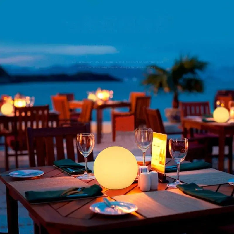 Gelb leuchtende Kugellampe als Tischdekoration auf einem gedeckten Restaurant-Terrassentisch mit Gläsern, Blick aufs abendliche Meer. 5