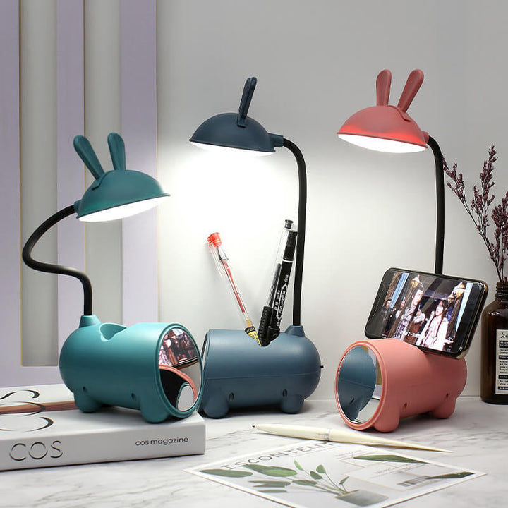 Drei leuchtende Hasenlampen in Grün, Dunkelblau und Rosa auf dem Schreibtisch, mit Stiften, Smartphone im Halter und Spiegel am Sockel. 1