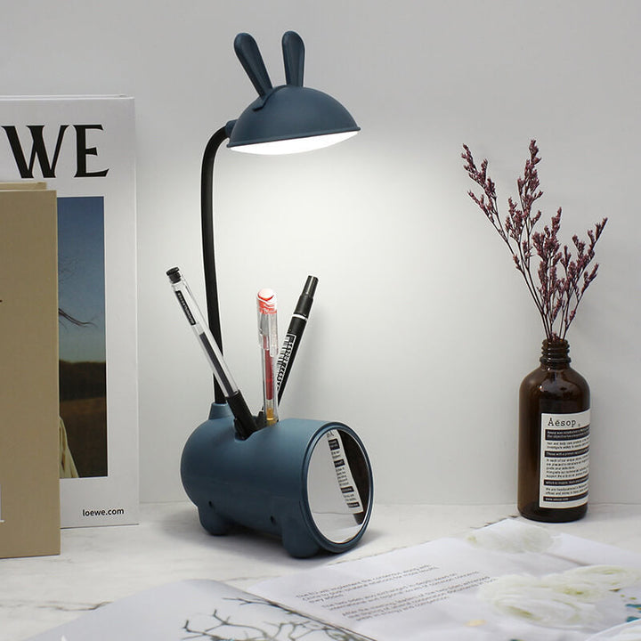 Dunkelblaue Hasenlampe leuchtet auf dem Schreibtisch, im Sockel stecken drei Stifte, daneben Zweige in einer Glasflasche und aufgeschlagene Bücher. 9