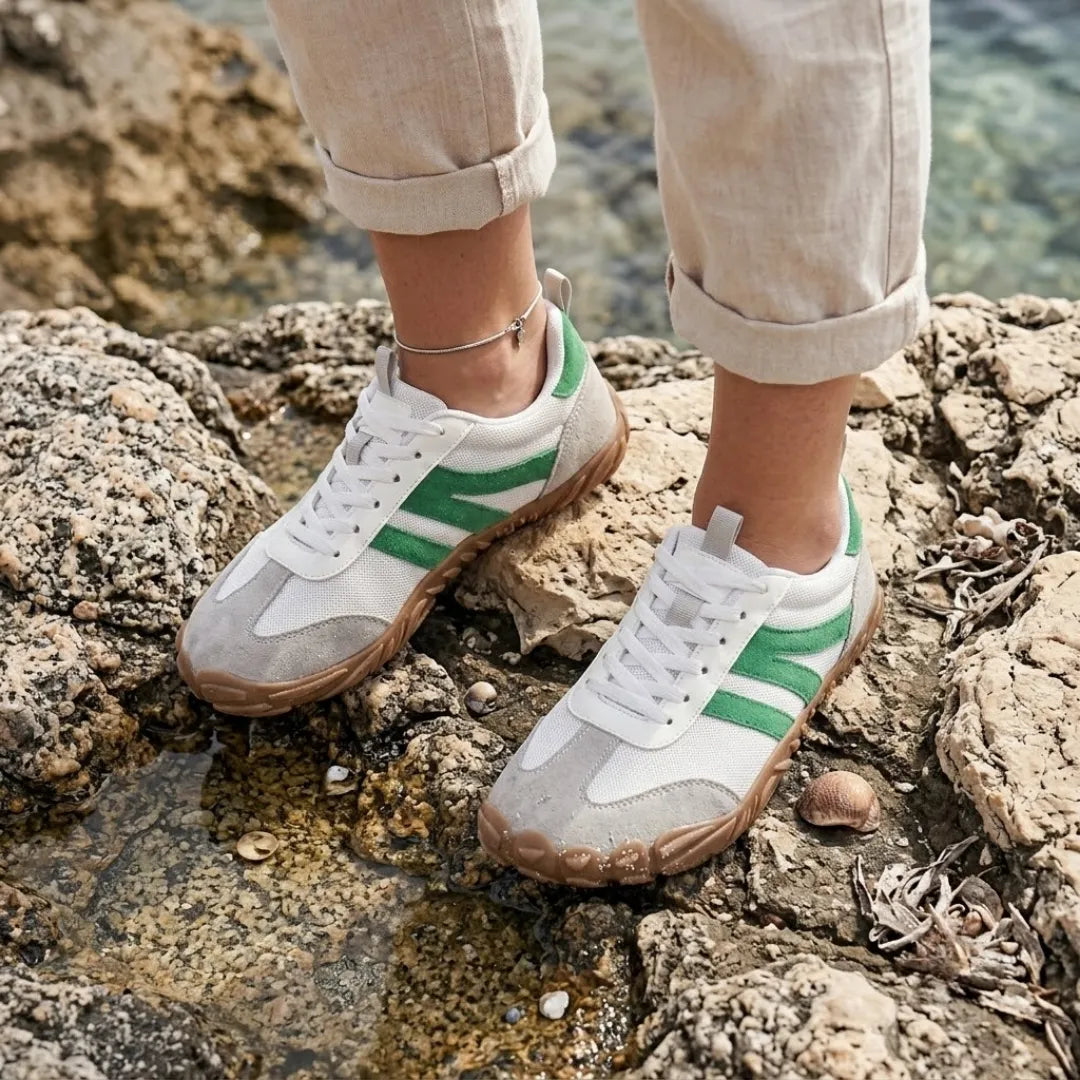 Frau trägt die weißen Barfußschuhe mit grünen Streifen, Leinenhose hochgekrempelt, auf Felsen am Wasser stehend. 29