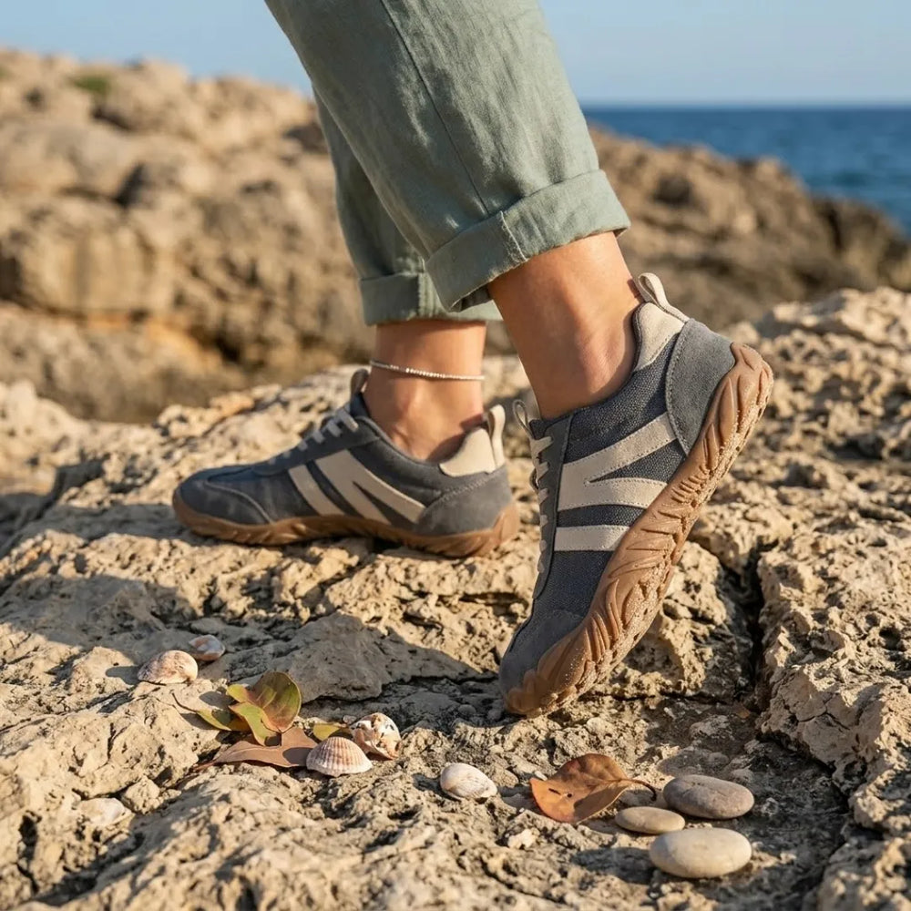 Graue Barfußschuhe an den Füßen einer Frau mit hochgekrempelter Hose, die auf Küstenfelsen mit Muscheln und Kieseln steht. 2