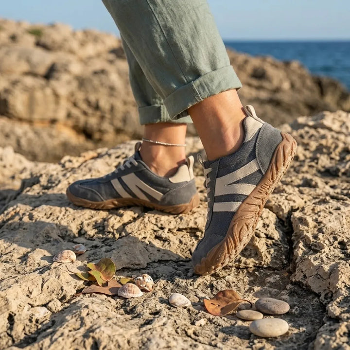 Graue Barfußschuhe an den Füßen einer Frau mit hochgekrempelter Hose, die auf Küstenfelsen mit Muscheln und Kieseln steht. 2