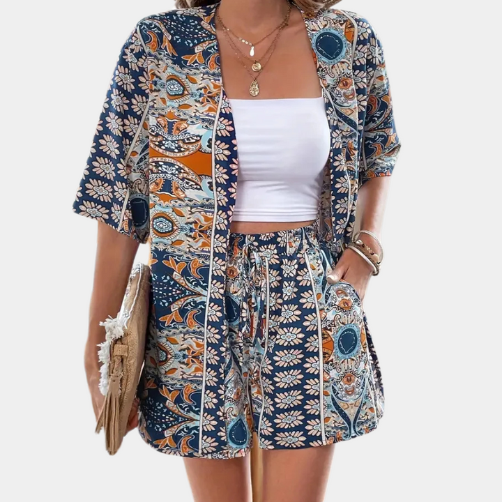 Frau in blau-orange gemustertem Sommeranzug, weißem Top, mit Strohtasche und Schmuck.