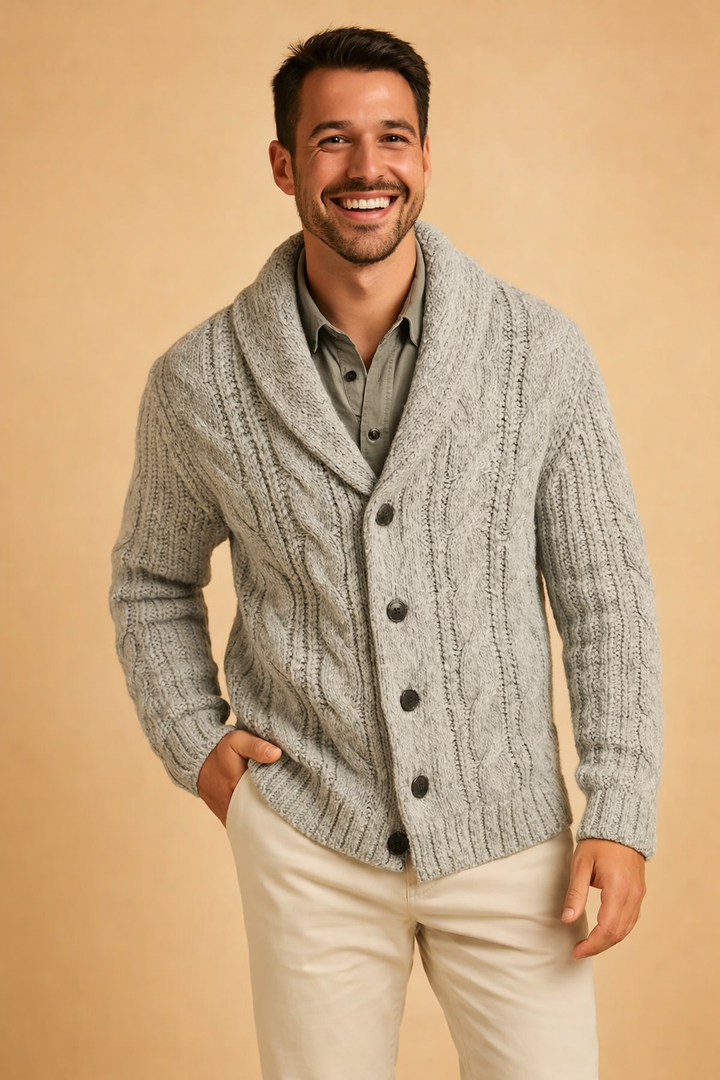 Lächelnder Mann in hellgrauer, grob gestrickter Strickjacke mit Schalkragen über olivgrünem Hemd, eine Hand in der Hosentasche. 7