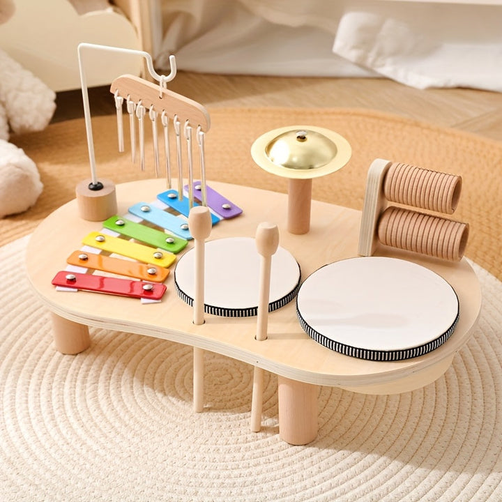 Holztisch mit Regenbogen-Xylophon, Klangstäben, Becken, zwei Trommeln und Rillenhölzern, Schlägel stecken in Halterungen, auf einem hellen Teppich. 1