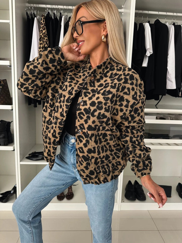 Frau in der Frontansicht trägt die Jacke mit Leopardenmuster offen über schwarzem Top und heller Jeans, eine Hand am Kopf. 1