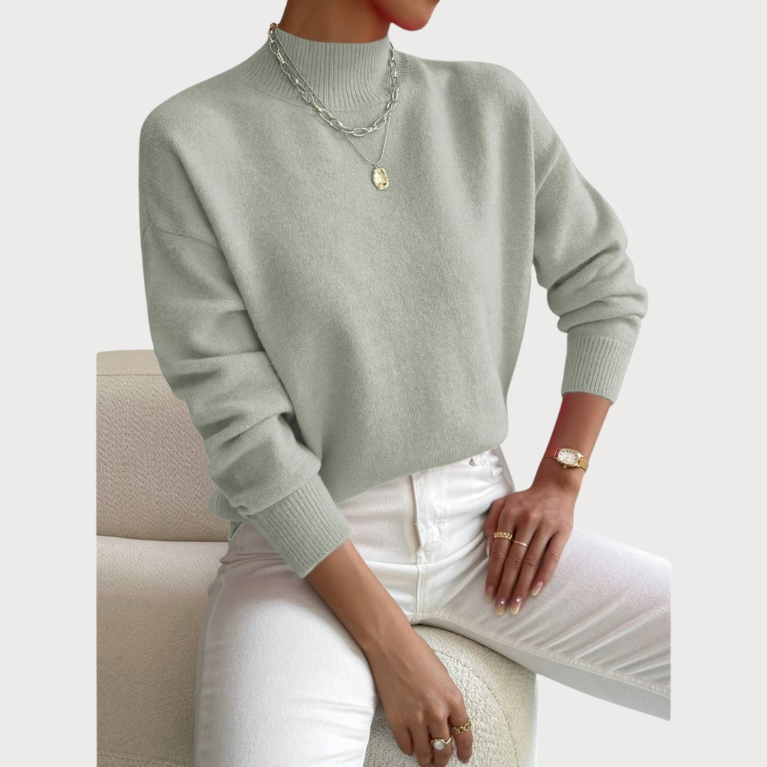 Salbeigrüner Pullover mit Stehkragen am Model, getragen mit weißer Jeans, goldenen Ketten und Armbanduhr auf einem hellen Bouclé-Sessel. 2