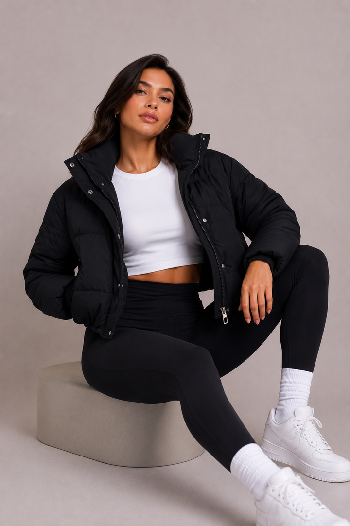 Model sitzt auf einem Podest und trägt die offene schwarze Steppjacke mit Stehkragen über weißem Crop-Top und schwarzer Leggings. 6