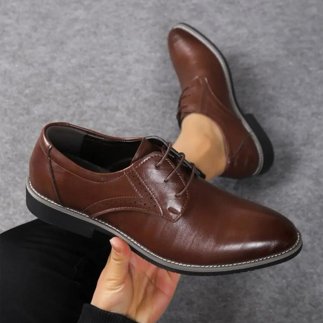 Dunkelbrauner Schnürschuh mit Zierlochung und heller Ziernaht in der Hand gehalten, daneben der zweite Schuh an einem Fuß. 2