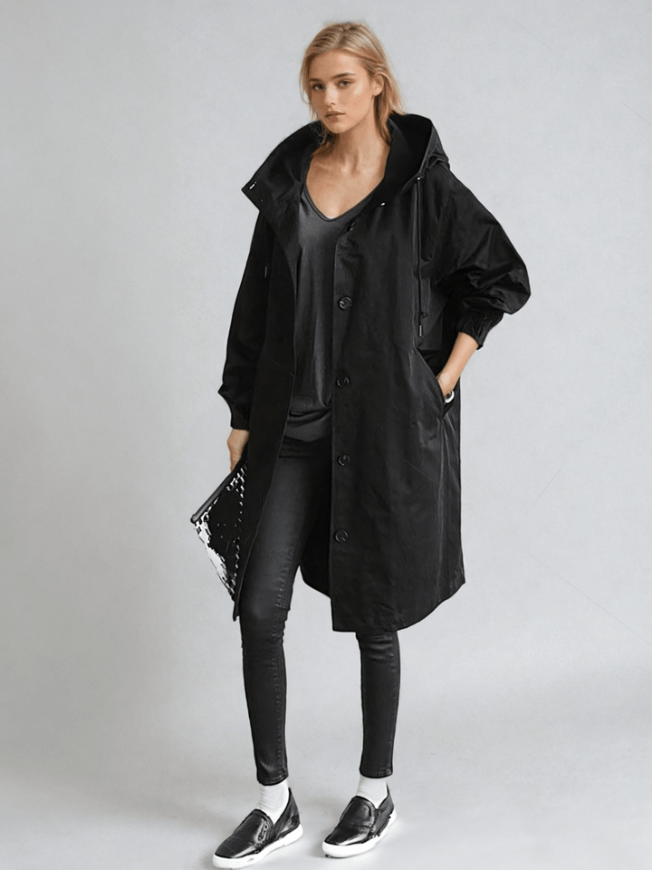 Ganzkörperansicht in Schwarz: Der Mantel mit Kapuze und Knopfleiste, kombiniert mit schwarzem Top, enger Hose, Slippern und Clutch. 2